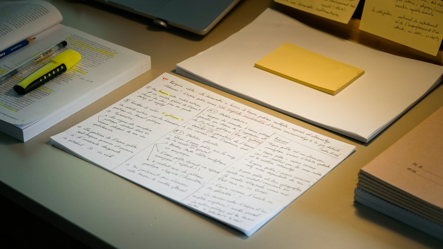 Studieboeken en handgeschreven notities verspreid over een rommelig bureau.